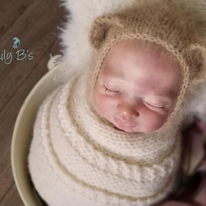 May include: A realistic newborn doll with closed eyes, wearing a tan fuzzy knit bonnet with small rounded ears. The doll is wrapped in a thick, cream-colored cable knit blanket. The text Lily B's is visible in the upper left corner of the image.