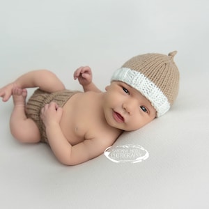 May include: A knitted tan and white beanie with a small top knot and matching tan knitted diaper cover. The beanie features a ribbed texture with a white band at the base, while the diaper cover has a ribbed waistband and leg openings.
