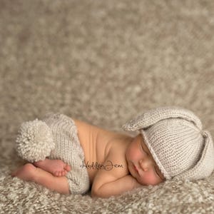 Op de afbeelding: Een beige gebreide outfit voor pasgeborenen bestaande uit een muts met lange konijnenoren en een bijpassende broek met een pluizige pomponstaart. De set is gemaakt van zacht, getextureerd garen en is ontworpen om op een konijn te lijken.