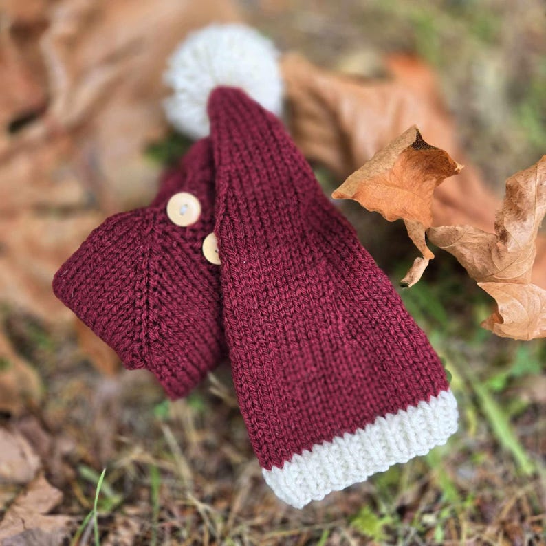 A rich, burgundy Santa Hat lays atop a knitted sweater in the same colour. There are a lot of dried fall leaves beneath it.