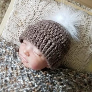 Puede incluir: Un gorro de ganchillo marr&oacute;n con un borde acanalado grueso y un pomp&oacute;n de piel sint&eacute;tica blanca en la parte superior. El gorro lo lleva una mu&ntilde;eca con los ojos cerrados, mostrando la construcci&oacute;n de hilo texturizado y el contraste de colores.