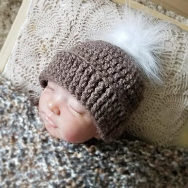 Puede incluir: Un gorro de ganchillo marr&oacute;n con un borde acanalado grueso y un pomp&oacute;n de piel sint&eacute;tica blanca en la parte superior. El gorro lo lleva una mu&ntilde;eca con los ojos cerrados, mostrando la construcci&oacute;n de hilo texturizado y el contraste de colores.