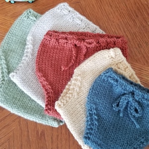 May include: A collection of five hand-knitted diaper covers in various solid colors including sage green, off-white, terracotta, cream, and dusty blue. Each piece features a ribbed waistband with a decorative knitted drawstring tie at the center.