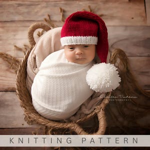 Pu&ograve; includere: Un berretto lavorato a maglia con corona rosso scuro, bordo a coste bianco e un grande pompon bianco sulla punta. Il berretto &egrave; indossato da un neonato avvolto in una coperta bianca in un cesto. Il testo KNITTING PATTERN &egrave; in basso.