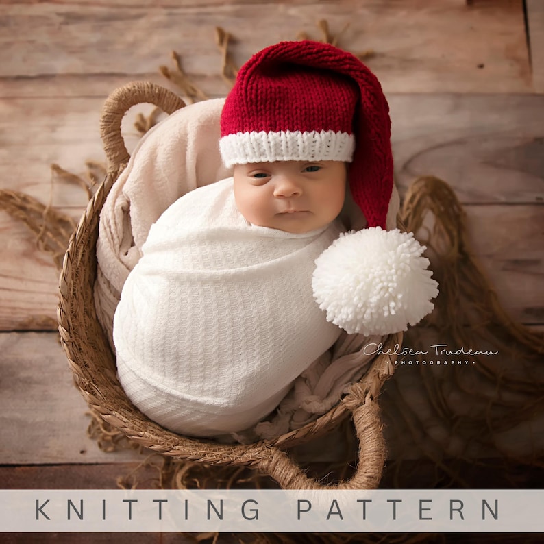 Pu&ograve; includere: Un berretto lavorato a maglia con corona rosso scuro, bordo a coste bianco e un grande pompon bianco sulla punta. Il berretto &egrave; indossato da un neonato avvolto in una coperta bianca in un cesto. Il testo KNITTING PATTERN &egrave; in basso.