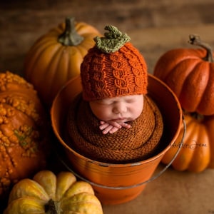 May include: A newborn infant wrapped in a brown knitted fabric, wearing a textured orange pumpkin-shaped hat with a green leaf accent, resting inside an orange metal bucket surrounded by several orange pumpkins of varying sizes and textures.