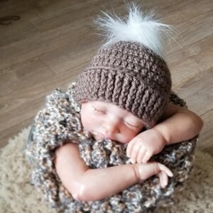 Puede incluir: Un gorro de ganchillo marr&oacute;n con borde acanalado y un pomp&oacute;n blanco esponjoso en la parte superior. El gorro lo lleva un beb&eacute; dormido envuelto en una manta de punto gruesa, texturizada y multicolor con tonos marrones, grises y blancos.