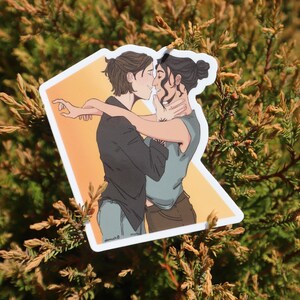 May include: A sticker featuring an illustration of two people embracing. One person has short brown hair and is wearing a black shirt and blue jeans. The other person has dark hair in a bun and is wearing a blue shirt and brown pants. The background is orange.