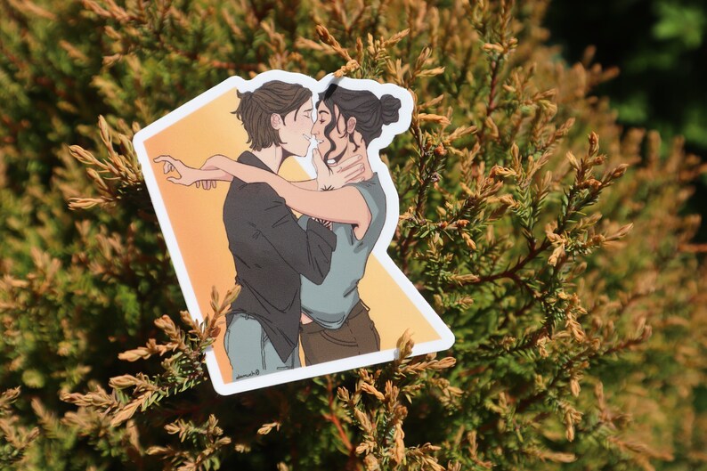 May include: A sticker featuring an illustration of two people embracing. One person has short brown hair and is wearing a black shirt and blue jeans. The other person has dark hair in a bun and is wearing a blue shirt and brown pants. The background is orange.