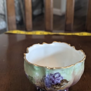 May include: A small, antique porcelain bowl with a scalloped rim and a floral design. The bowl features a white interior, a gold-painted rim, and a painted exterior with green, purple, and brown hues. The bowl is likely used for serving or display.