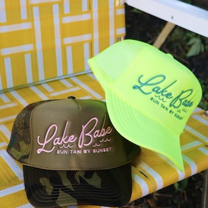 Könnte beinhalten: Zwei Baseballkappen mit dem Text "Lake Babe" werden auf einem gelb-weißen Stuhl gezeigt. Eine Kappe ist olivgrün mit rosa Schriftzug und einem Camouflage-Schirm. Die andere ist neongrün mit blauem Schriftzug.