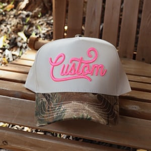 May include: A beige baseball cap with a camouflage brim. The word "Custom" is embroidered in bright pink cursive on the front of the hat. The hat is sitting on a wooden bench.