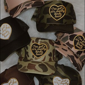 May include: A collection of six camouflage trucker hats with a heart-shaped patch that says "I've got a 90's country kinda heart". The hats are in various shades of green, brown, and beige.