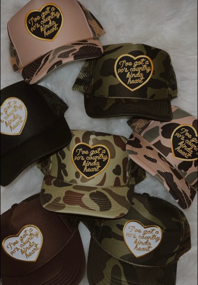 May include: A collection of six camouflage trucker hats with a heart-shaped patch that says "I've got a 90's country kinda heart". The hats are in various shades of green, brown, and beige.