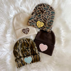 Pode incluir: Quatro gorros de malha com padrões e cores diferentes. Cada gorro tem um remendo em forma de coração com o texto "I've got a 90's country kinda heart". Os remendos são de diferentes cores: amarelo, castanho, rosa e azul.
