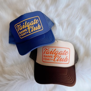 Może przedstawiać: Dwie czapki trucker z napisem "Tailgate Club Game Day" w retro stylu. Czapka po lewej stronie jest niebieska z pomarańczowym napisem, a czapka po prawej stronie jest beżowa z pomarańczowym napisem.