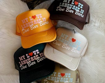 May include: Six trucker hats with a mesh back in different colors and patterns. Each hat has the same text: "My Love Language is Dive Bar Beers" with a pink heart in the text.
