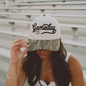 Op de afbeelding: Een witte baseballcap met de tekst "Gameday" in zwart en een football-afbeelding. De klep van de cap heeft een camouflagepatroon. De cap wordt gedragen.