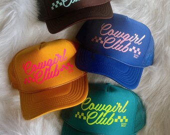 May include: Four trucker hats with a "Cowgirl Club" logo in different colors. The hats are brown, blue, yellow, and green. The logo is in a cursive font with a checkered pattern.