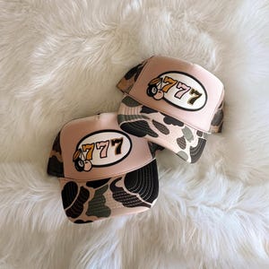 May include: Two pink and camo trucker hats with a white oval patch that says "777" and a cartoon cherry and 8-ball graphic.