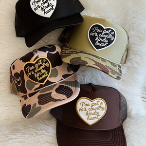 May include: Four trucker hats with a heart-shaped patch that says "I've got a 90's country kinda heart". The hats are in black, brown, camouflage, and green camouflage.