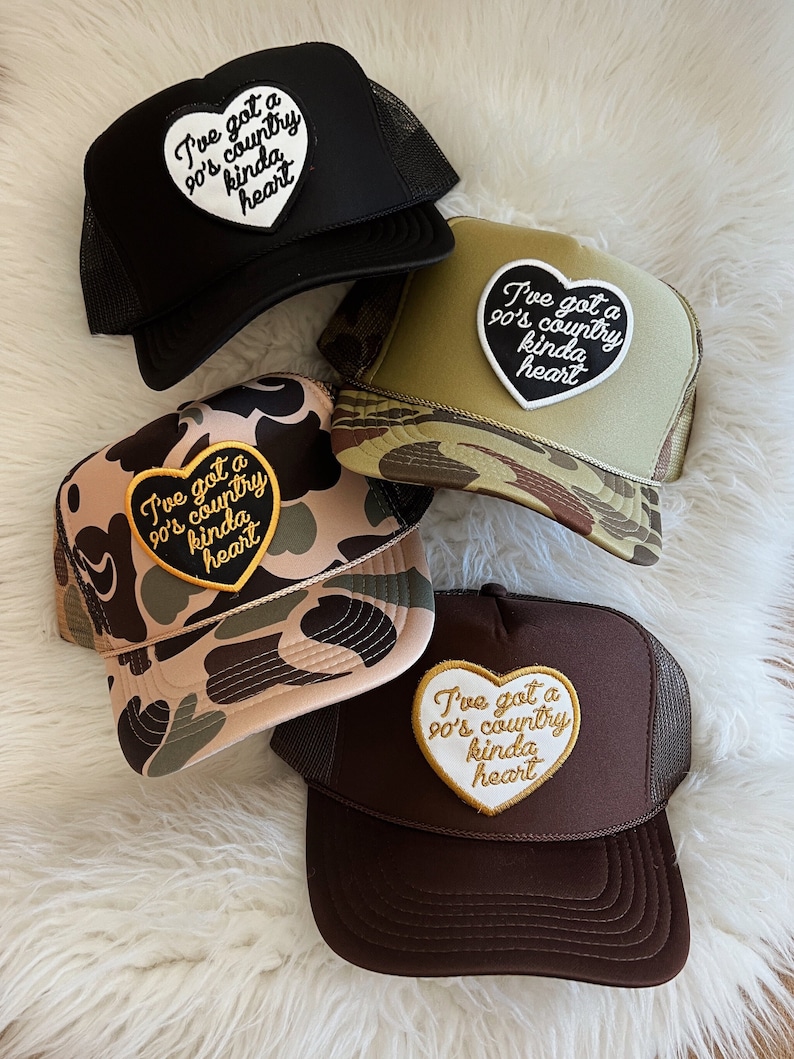 May include: Four trucker hats with a heart-shaped patch that says "I've got a 90's country kinda heart". The hats are in black, brown, camouflage, and green camouflage.