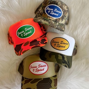 Op de afbeelding: Vier trucker caps met camouflagepatronen in verschillende kleuren. Elke cap heeft een patch met de tekst "Jesus Saves I Spend" in witte letters op een gekleurde achtergrond.