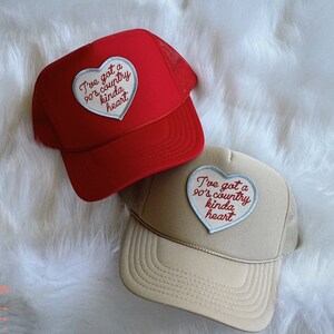 May include: Two trucker hats with a heart-shaped patch that says "I've got a 90's country kinda heart". One hat is red and the other is beige.