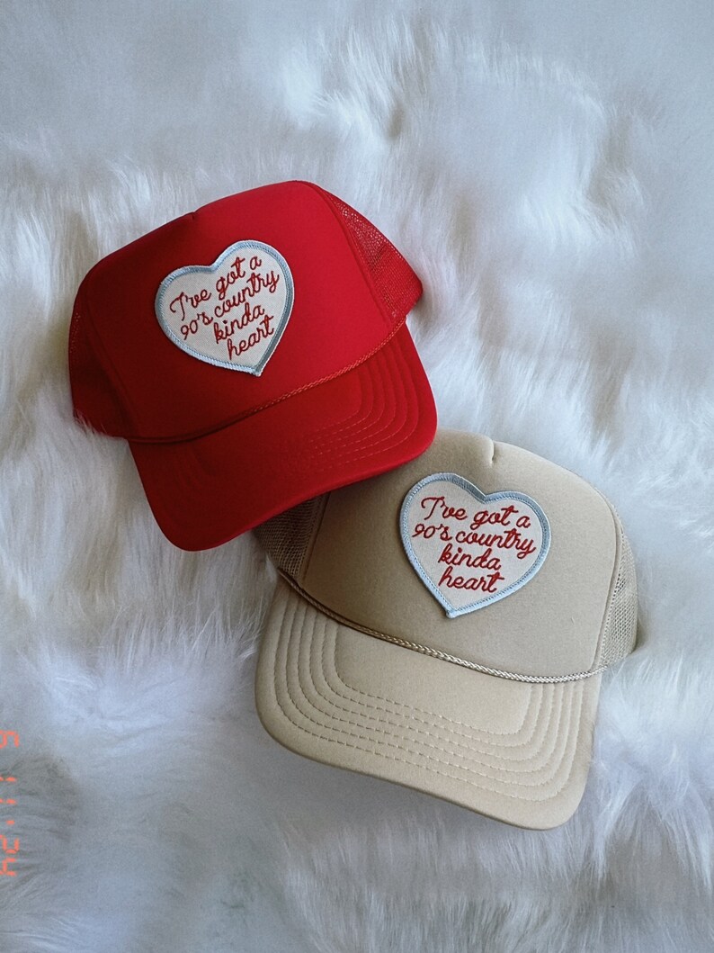 May include: Two trucker hats with a heart-shaped patch that says "I've got a 90's country kinda heart". One hat is red and the other is beige.