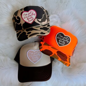 May include: Three trucker hats with a camouflage, orange, and beige design. Each hat features a heart-shaped patch with the text "I've got a 90's country kinda heart".