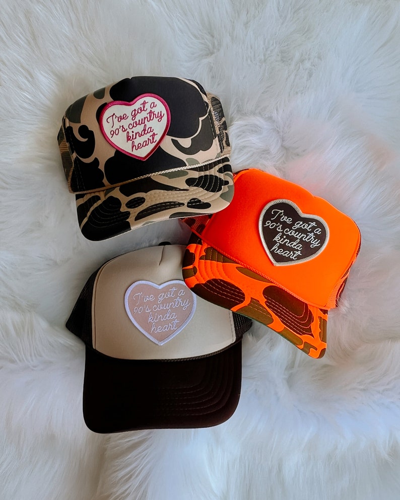 May include: Three trucker hats with a camouflage, orange, and beige design. Each hat features a heart-shaped patch with the text "I've got a 90's country kinda heart".