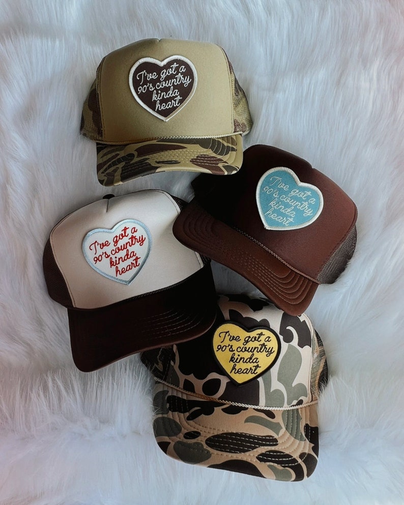 May include: Four trucker hats with a heart-shaped patch that says "I've got a 90's country kinda heart". The patches are in different colours: blue, red, yellow, and white. The hats are in different colours: brown, beige, and camouflage.