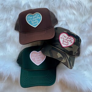 May include: Three trucker hats with different coloured mesh backs and embroidered patches. The patches are pink, blue, and white with the text "I've got a 90's country kinda heart".