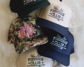 Puede incluir: Varias gorras de béisbol en diversos colores, como verde, azul marino y camuflaje. Cada gorra presenta el texto "Wilder Made FISHING CLUB" con un diseño de caña de pescar. Las gorras están dispuestas sobre una superficie blanca y esponjosa.