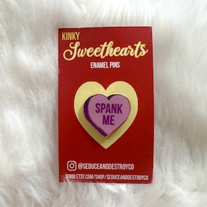 May include: A pink and purple enamel pin shaped like a conversation heart with the text "SPANK ME" in yellow letters. The pin is on a red card with the text "Kinky Sweethearts Enamel Pins" and the social media handle "@seduceanddestroyco".