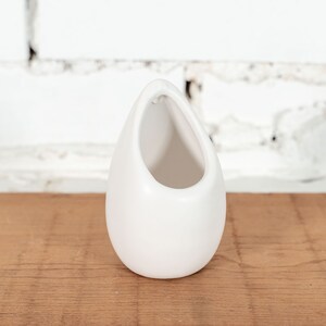 May include: White ceramic planter shaped like a teardrop with a wide opening at the top. The planter is sitting on a wooden surface.
