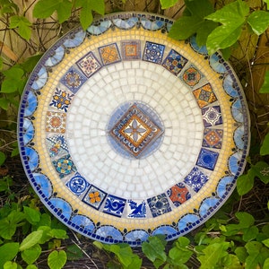 May include: A round mosaic table top with a white center and a blue and yellow border. The mosaic tiles are arranged in a circular pattern and feature various designs.