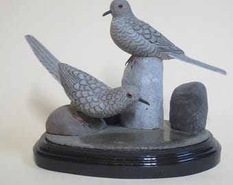 May include: A hand-carved wooden sculpture of two mourning doves perched on rocks. The doves are painted in shades of gray and have realistic details. The sculpture is displayed on a black base.
