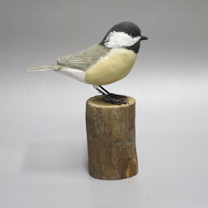 May include: A hand-carved wooden bird sculpture of a chickadee perched on a wooden log. The bird is painted in shades of gray, white, and black, with realistic details.