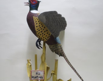 May include: A colorful pheasant sculpture with a detailed wing and tail feathers, perched on a corn stalk base. The bird features red, blue, yellow, and brown hues. A small sign reads "NO HUNTING".