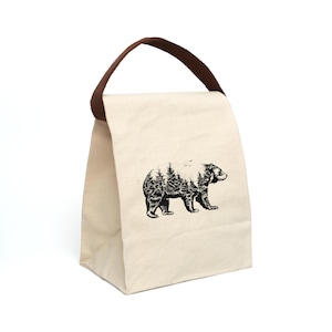 May include: A white canvas lunch bag with a brown handle. The bag features a black and white illustration of a bear with a forest inside its silhouette.