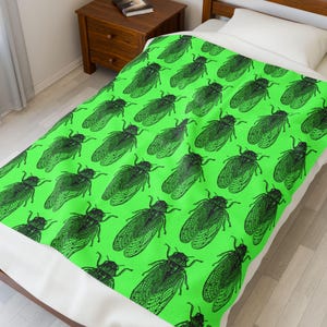Op de afbeelding: Een fel limoengroene deken met een herhaald patroon van zwarte cicade-illustraties. De deken ligt op een bed met een wit kussen en een houten nachtkastje. De cicaden zijn gedetailleerd met zichtbare vleugels en lichaamssegmenten.