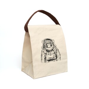 May include: A white canvas lunch bag with brown handles. The bag features a black and white illustration of a chimpanzee wearing an astronaut suit.