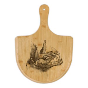 Puede incluir: Una tabla de cortar de bambú con asa, con una ilustración grabada de la cabeza de un murciélago gruñendo. La cara del murciélago está intrincadamente dibujada con dientes afilados y orejas puntiagudas. La tabla tiene una veta de madera natural.
