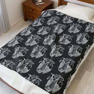 May include: A black and white patterned blanket featuring a repeating design of bats. The bats are depicted in a detailed, vintage style, with their wings spread. The blanket is on a bed with a white pillow and a wooden nightstand.