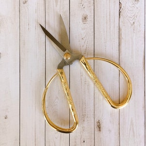 May include: Gold-colored scissors with ornate handles against a woodgrain background.