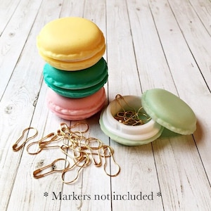 May include: A set of three pastel-colored macaron-shaped containers with a single green macaron container open and filled with gold safety pins. The text "*Markers not included*" is written on the wooden surface below the containers.