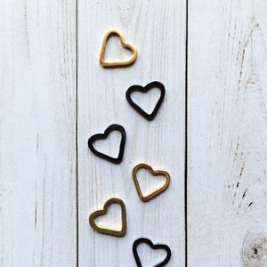 May include: Six heart-shaped metal charms, three gold and three black, arranged vertically on a white wooden background.
