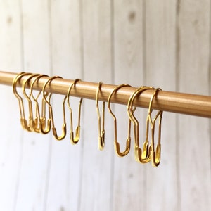 May include: A set of gold-toned metal shower curtain rings hanging on a wooden rod. The rings are in the shape of a safety pin.
