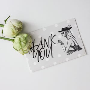 May include: A gray card with white polka dots and the words "THANK YOU" in black script. An illustration of a woman in a hat and sunglasses is on the card. Two white and green roses are on the left.